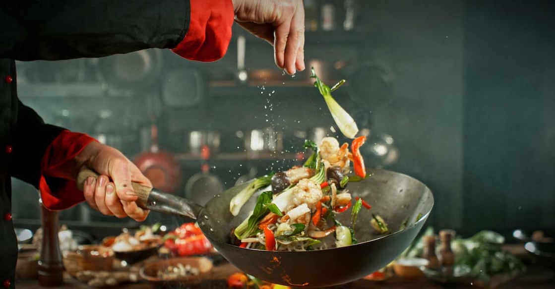 2185198074 Freeze Motion of Wok Pan with Flying Ingredients in the Air. Chef Pouring Salt. Kitchen on Background.