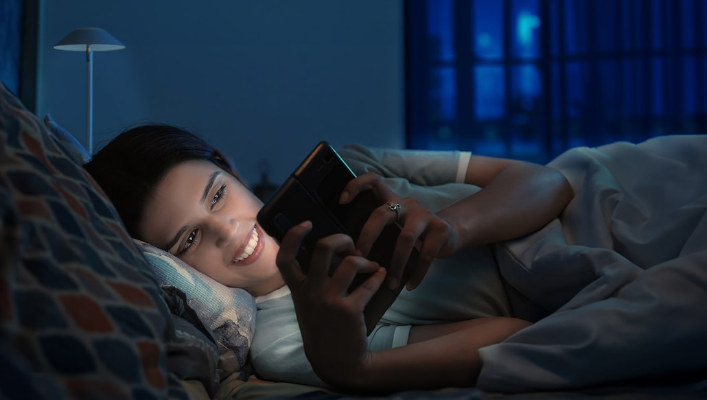 2130055944 Beautiful Indian young adult girl lying on bed using mobile phone enjoy chatting late night at indoor home. Happy smiling adult woman looking on smartphone screen typing reading massage news at inside house