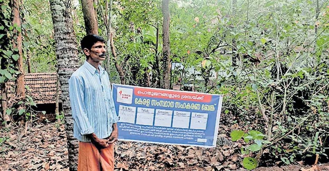 muttanur-airport-land-money-issue കാനാട് നല്ലാണിയിലെ വീടിന് മുന്നിൽ ജപ്തി വിവരം കാണിച്ച് കേരള ബാങ്ക് സ്ഥാപിച്ച ബാനറിനു മുന്നിൽ നിൽക്കുന്ന കെ.സനിൽ