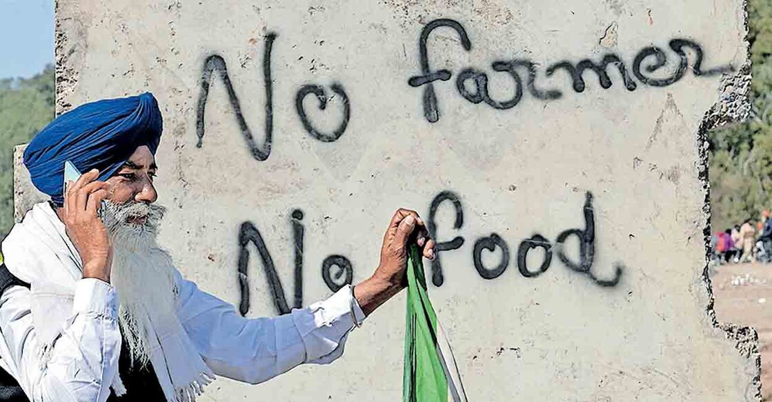 farmers-protest പഞ്ചാബ് – ഹരിയാന അതിർത്തിയിലെ ശംഭുവിൽ ‘കർഷകരില്ലെങ്കിൽ ഭക്ഷണമില്ല’ എന്നെഴുതിവച്ച ബോർഡിനു മുന്നിൽ സമരക്കാരിലൊരാൾ ഫോണിൽ സംസാരിക്കുന്നു. ചിത്രം: എഎഫ്പി