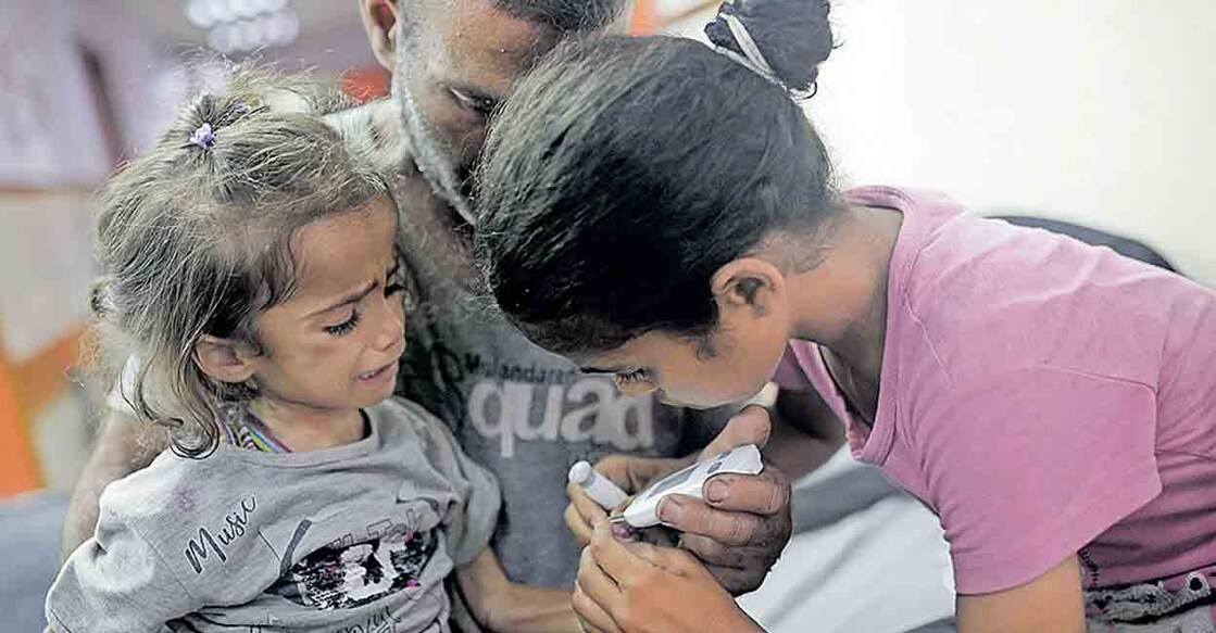 യുദ്ധത്താൽ പോഷകാഹാരം ലഭിക്കാതെ വന്നതു മൂലം ആരോഗ്യക്കുറവ് അനുഭവപ്പെടുന്ന ഹന അൽ റായിയുടെ പ്രമേഹനില സഹോദരി പരിശോധിക്കുന്നു. മൂന്നുവയസ്സുകാരിയായ ഹനയെ എടുത്തിരിക്കുന്നത് പിതാവ് അബ്ദുൽ റഹ്മാനാണ്. അകാല പ്രമേഹം, ദുർബലമായ പ്രതിരോധ വ്യവസ്ഥ എന്നിവ ഹനയ്ക്കുണ്ട്. ഗാസയിലെ ദൈർ അൽ ബലായിലെ ആശുപത്രിയിൽ നിന്നുള്ള ദൃശ്യം. വടക്കൻ ഗാസയിൽ നിന്നു പലായനം ചെയ്തതാണ് ഹനയും കുടുംബവും. ചിത്രം: എപി 