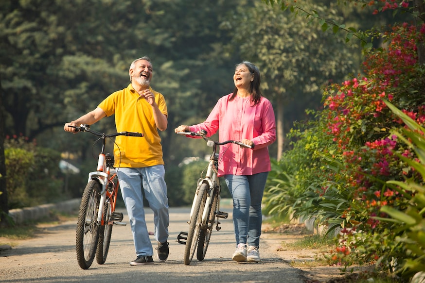 1448553779 Carefree senior couple talking and laughing while walking with bicycles on road amidst plants in park during weekend