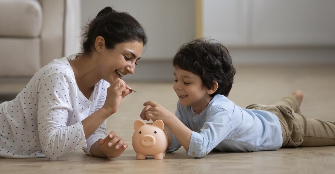 Mother and Child Caring young asian indian mother teaching little kid son saving money or planning future purchases, putting coins in piggybank, lying on heated floor, financial education for children concept.