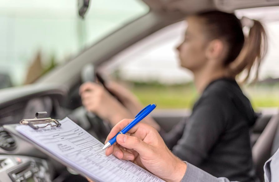 697993984 Teenage Girl Taking A Driving Lesson during bright sunny day. Closeup of a instructor man writing on a clipboard and taking notes while sitting next to teen driver.
