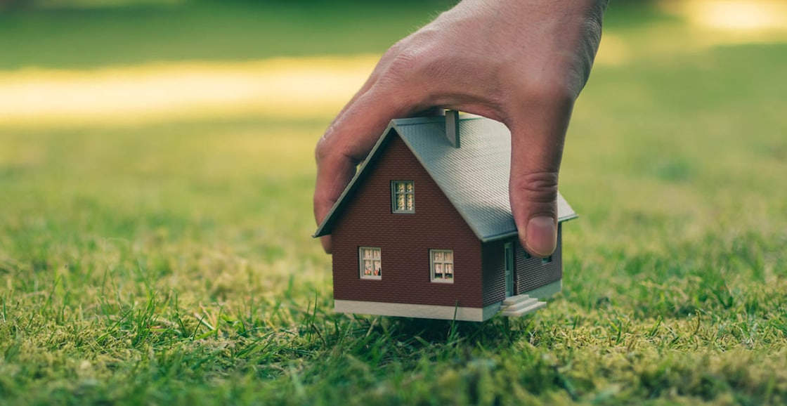 Concept of selling a house. A hand is holding a model house above green meadow.