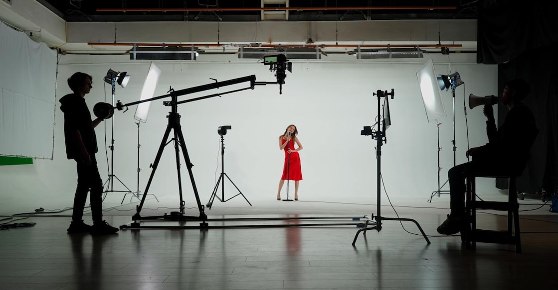 Professional filming pavilion with a white cyclorama. Shooting of a girl in a red dress who sings into a retro microphone. Director, Cameraman and crew in Backstage.