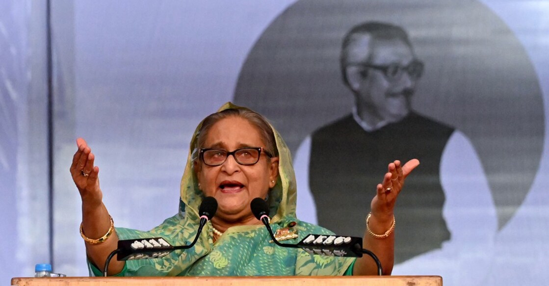 sheikh-hasina-background-mujibur-rahman 2023 ഡിസംബറിൽ ഒരു തിരഞ്ഞെടുപ്പ് റാലിയെ അഭിസംബോധന ചെയ്തു സംസാരിക്കുന്ന ബംഗ്ലദേശ് പ്രധാനമന്ത്രി ഷെയ്ഖ് ഹസീന. പിതാവും രാഷ്ട്രപിതാവുമായ മുജിബുർ റഹ്മാന്റെ ചിത്രം പശ്ചാത്തലത്തിൽ (File Photo by AFP)