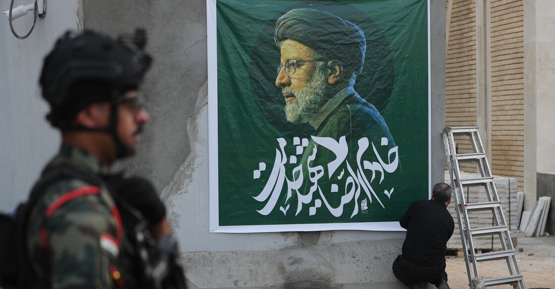 A member of the Iraqi security forces stands guard outside the Iranian embassy in Baghdad as a man hangs a huge portrait of Iran's late president Ebrahim Raisi at the entrance during a condolences service on May 20, 2024 for the president and his entourage, who were killed in a helicopter crash in Iran the previous day. (Photo by AHMAD AL-RUBAYE / AFP)