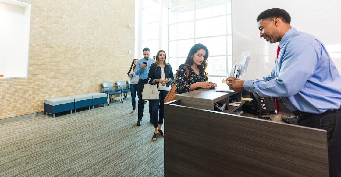 As a line of diverse people wait patiently, the mature adult male banker counts out the money that young adult woman has withdrawn from her savings account.