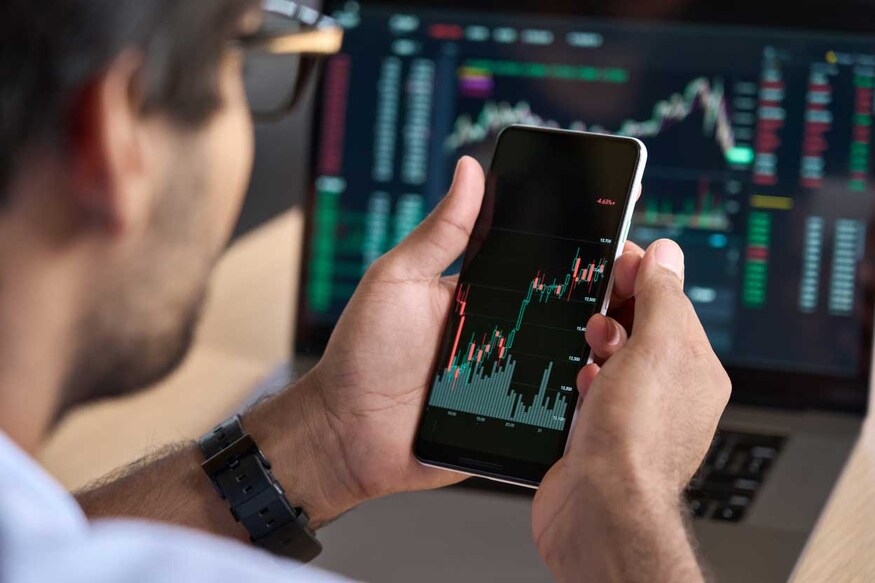 2111976929 Male trader investor broker crypto analyst holding smartphone in hand analyzing stock market trading charts indexes data checking price using mobile stockmarket exchange app, over shoulder view.