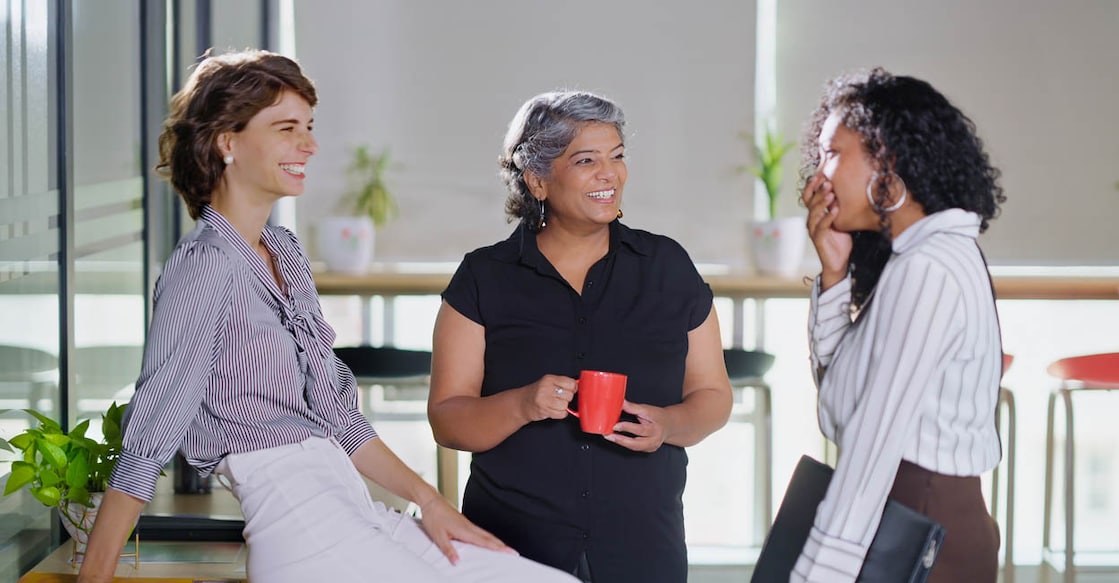 Indian smiling old business women standing hold cup drink coffee talk gossip indoor work place. Beautiful young adult corporate girls make funny blab share ideas during tea break spend time job office