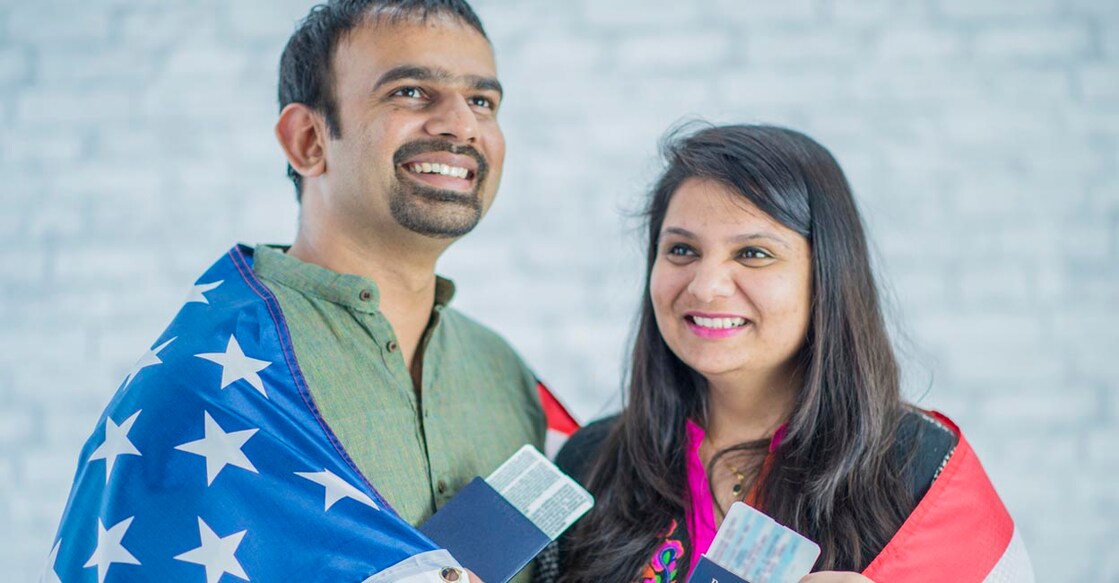 An Indian husband and wife are indoors in a new home. They are wearing Indian-style clothing. They are posing with their American passports and flight tickets, and they have an American flag draped around them.