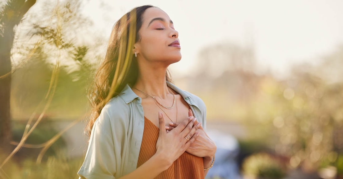 1496615471 Young latin woman with hand on chest breathing in fresh air in a beautiful garden during sunset. Healthy mexican girl enjoying nature while meditating during morning exercise routine with closed eyes. Mindfulness woman enjoying morning ritual and relaxing technique.